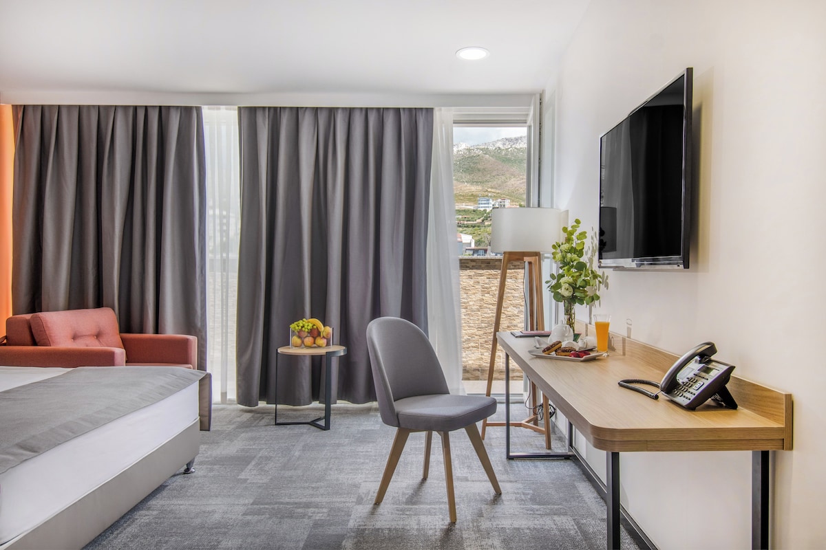 A hotel room with a bed, a desk with a telephone, a chair, a small table with fruit, and a TV on the wall.