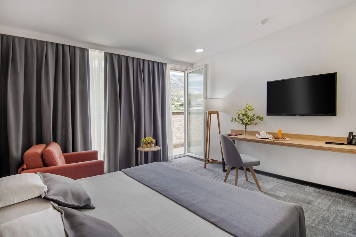 Habitación de hotel con cama, escritorio con televisor de pantalla plana, una silla roja, una mesita con frutas y vistas al exterior.