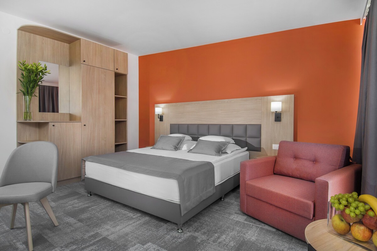 Quarto de hotel moderno com uma cama, uma escrivaninha, uma cadeira e uma pequena mesa com frutas.