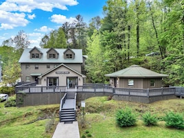 Um edifício de dois andares com uma sala de lareira e uma estrutura separada com telhado verde, cercado por uma cerca de madeira e vegetação exuberante.