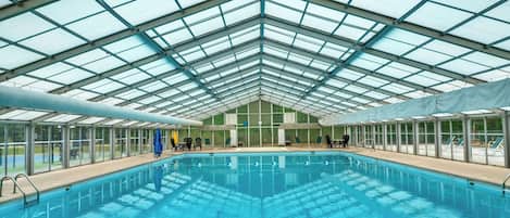 An indoor swimming pool with a glass ceiling, surrounded by lounge chairs and tables.