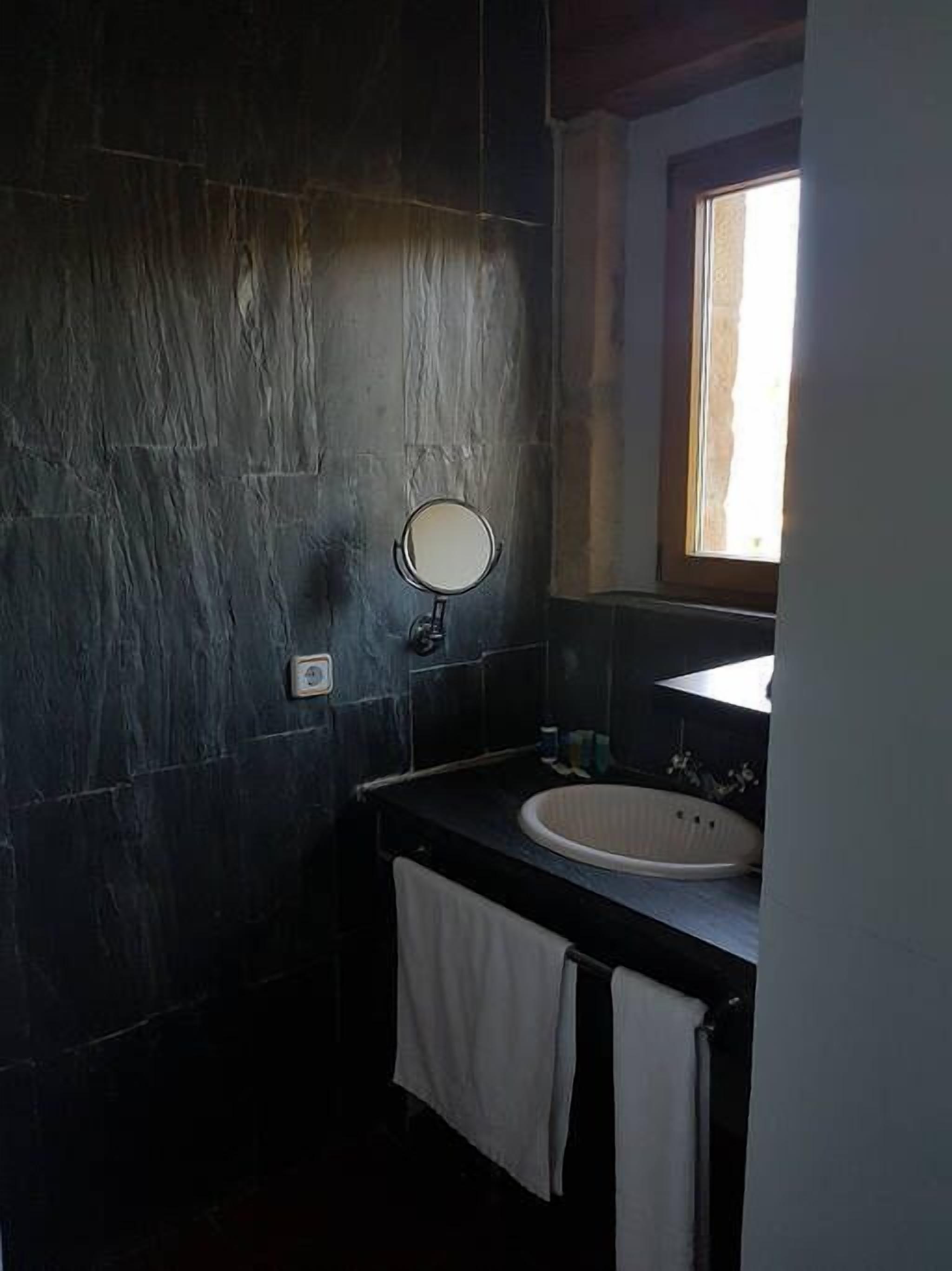 Un baño con paredes de piedra oscura, un espejo redondo, un lavamanos y una ventana.