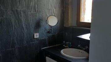 Un baño con paredes de piedra oscura, un espejo redondo, un lavamanos y una ventana.