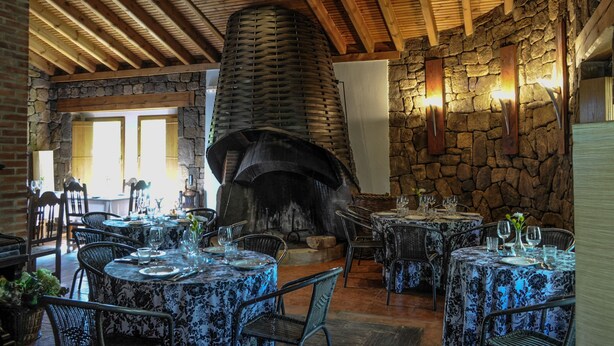 Un comedor rústico con paredes de piedra, vigas de madera y una gran chimenea. Las mesas están dispuestas con copas y platos.