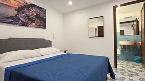 Un dormitorio con cama, una mesita de noche, un cuadro, un espejo y un baño visible a través de una puerta abierta.