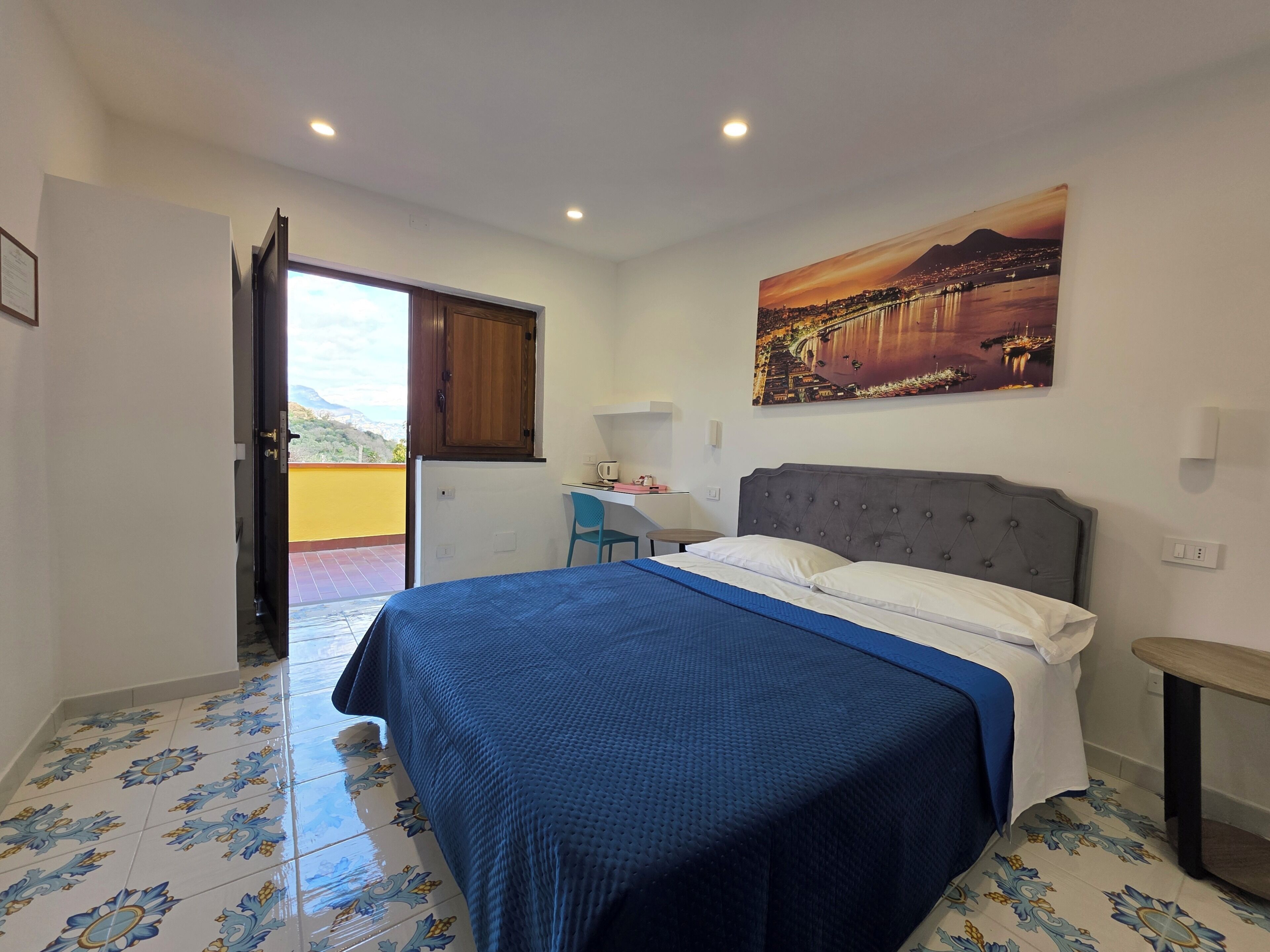 A bedroom with a bed, a desk, a chair, a painting, and a door leading to a balcony.