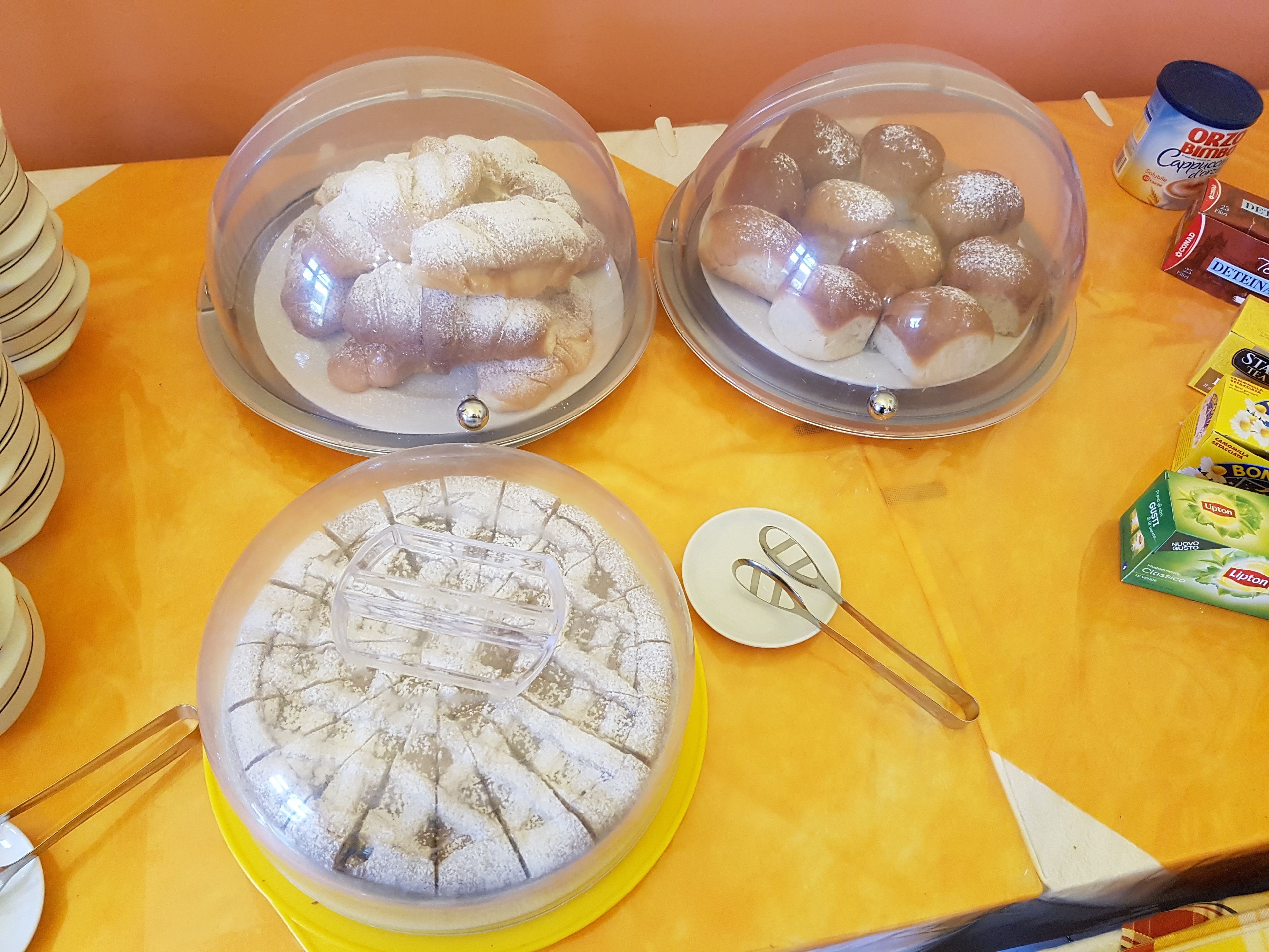 Ein reichhaltiges Buffet mit abgedeckten Schalen und Behältern, darunter Kuchen, Torten und verpackte Snacks.