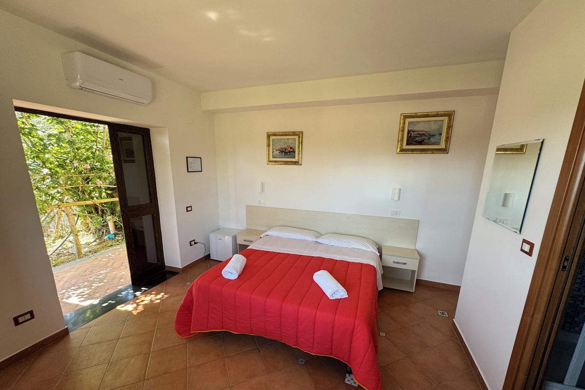 Un dormitorio con cama, mesitas de noche, un escritorio pequeño, un espejo y una puerta que da a un balcón con plantas.