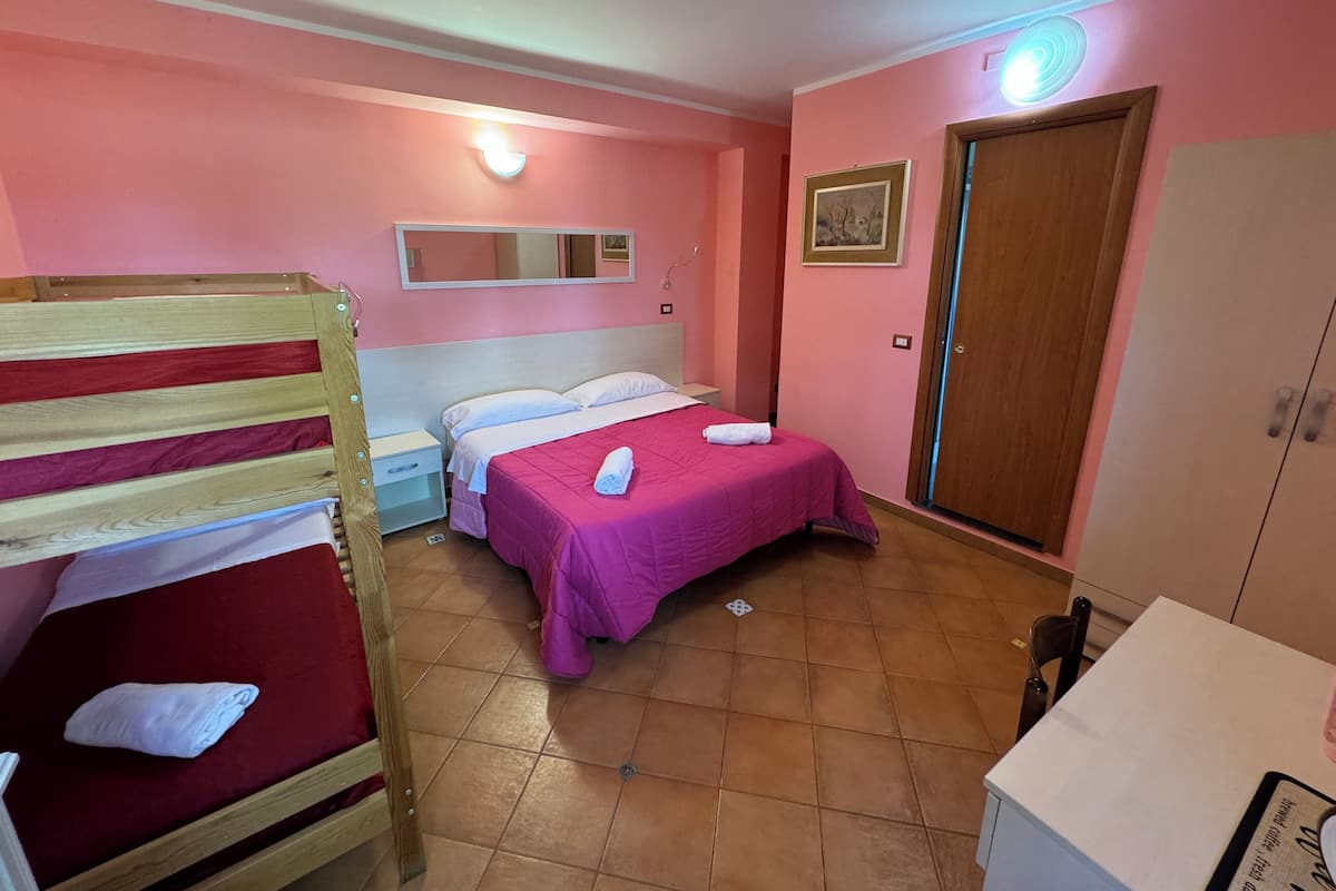 Una habitación con una cama litera, una cama individual con colcha rosa, una mesita de noche, un armario de madera y una puerta con luz encima.