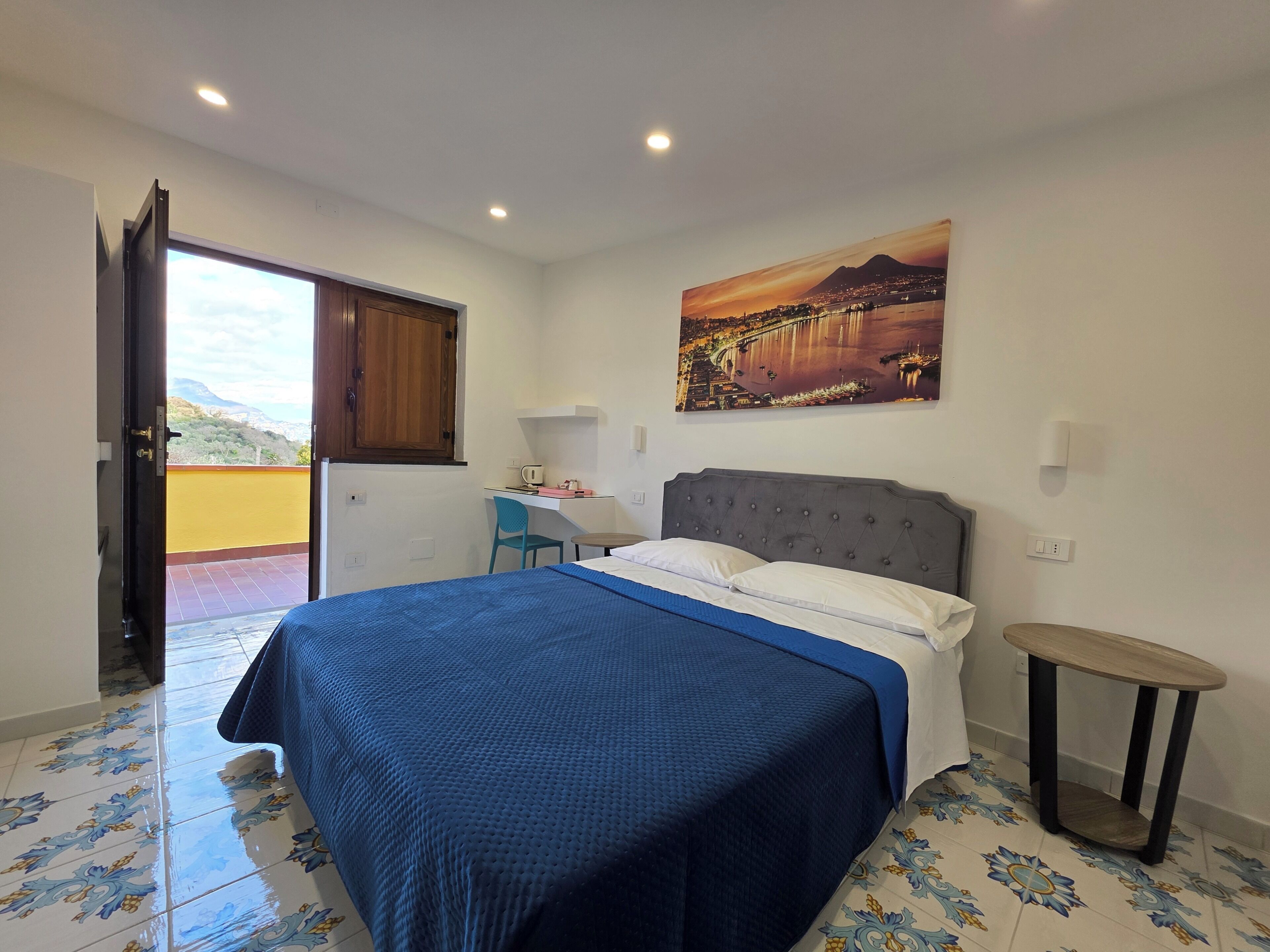 A bedroom with a bed, a desk, a chair, a small table, and a painting on the wall.