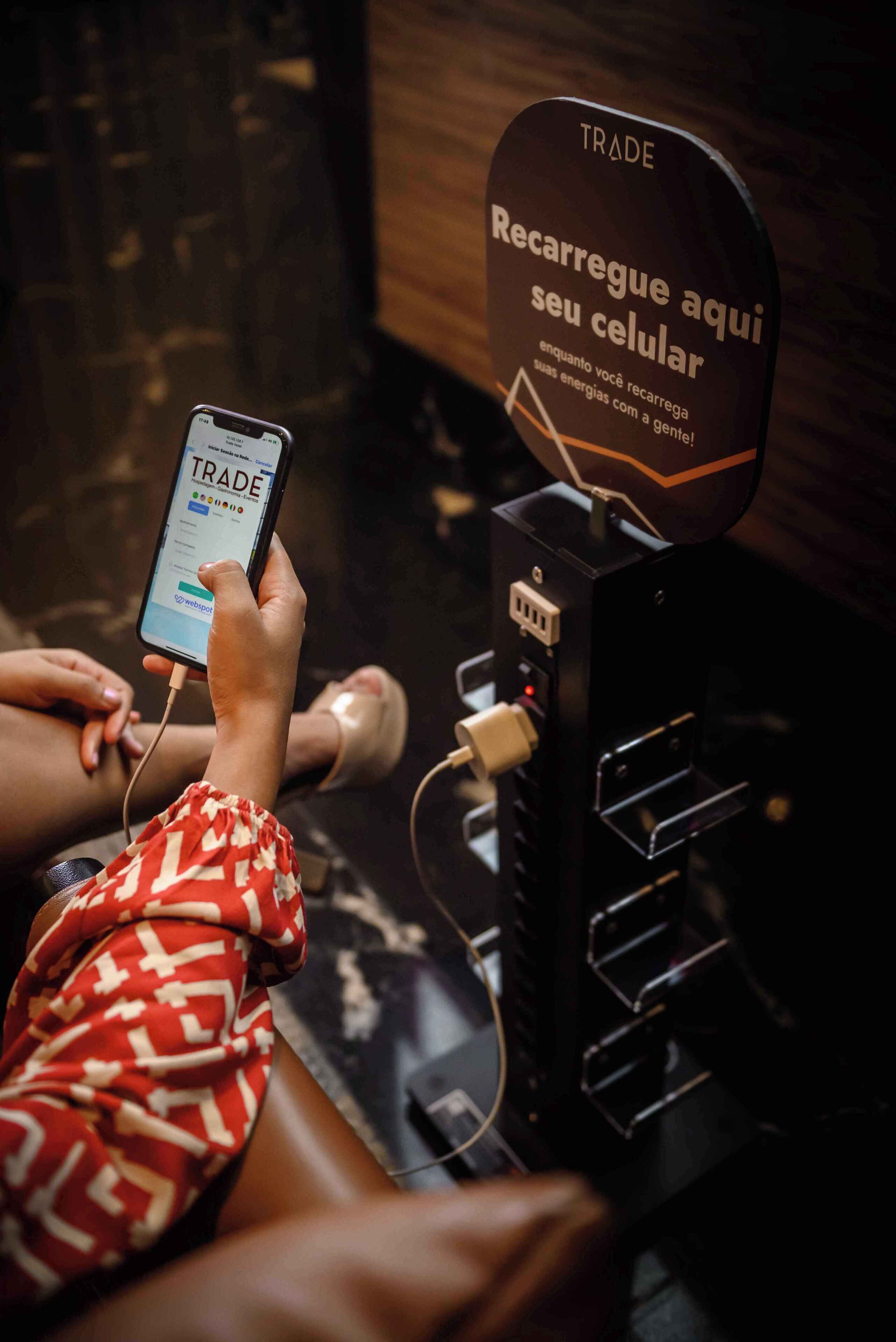 A person using a mobile device at a charging station with a sign that reads "Trade Recharge your cellphone here."
