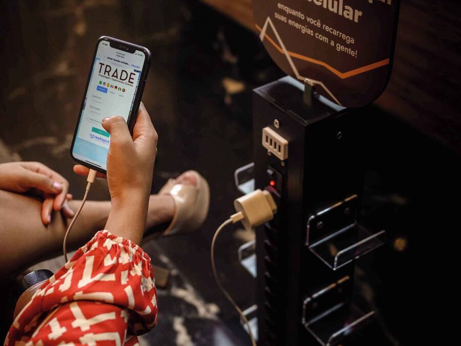 A person using a mobile device at a charging station with a sign that reads "Trade Recharge your cellphone here."