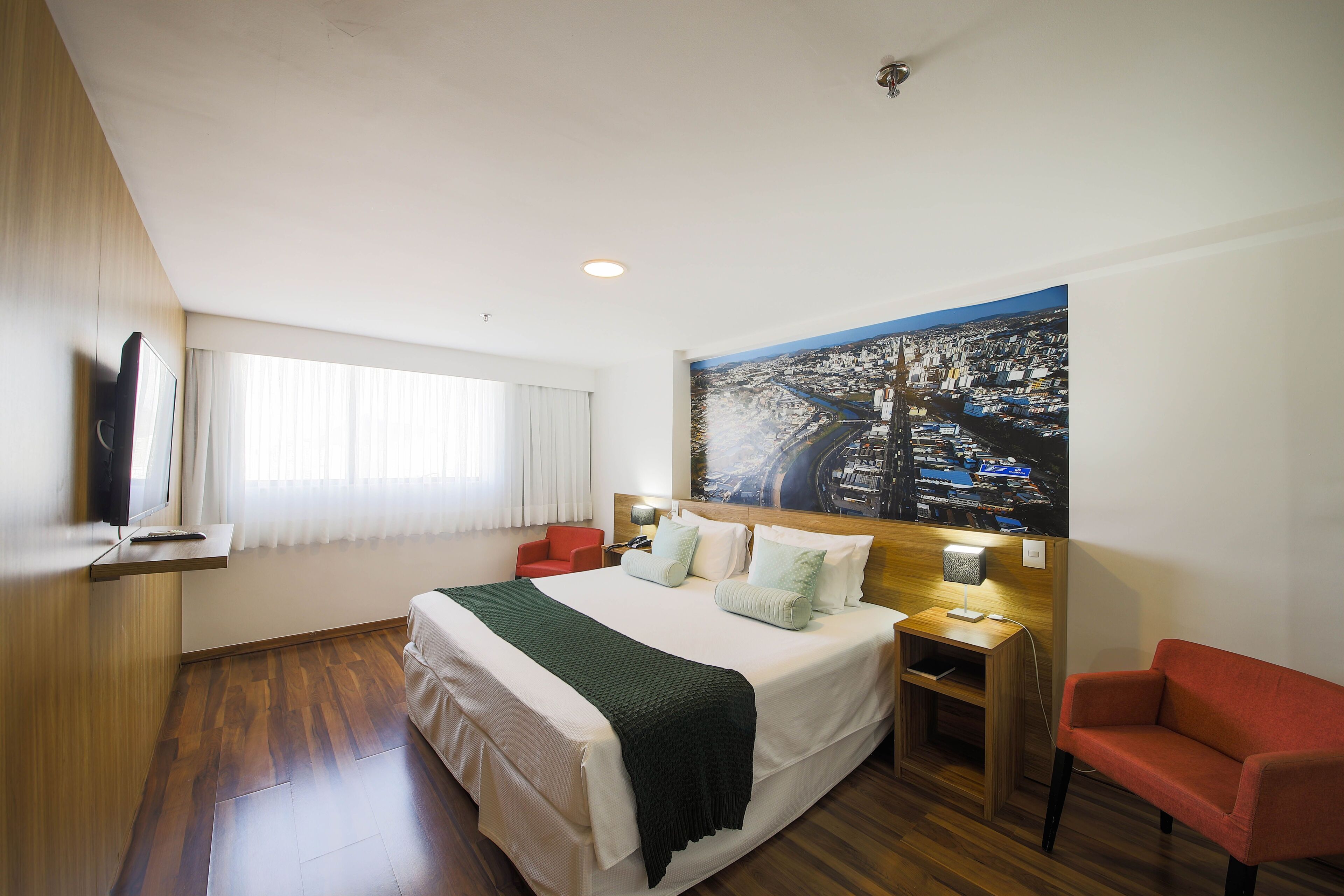 A hotel room with a large bed, a flat-screen TV, a red armchair, and a cityscape painting on the wall.