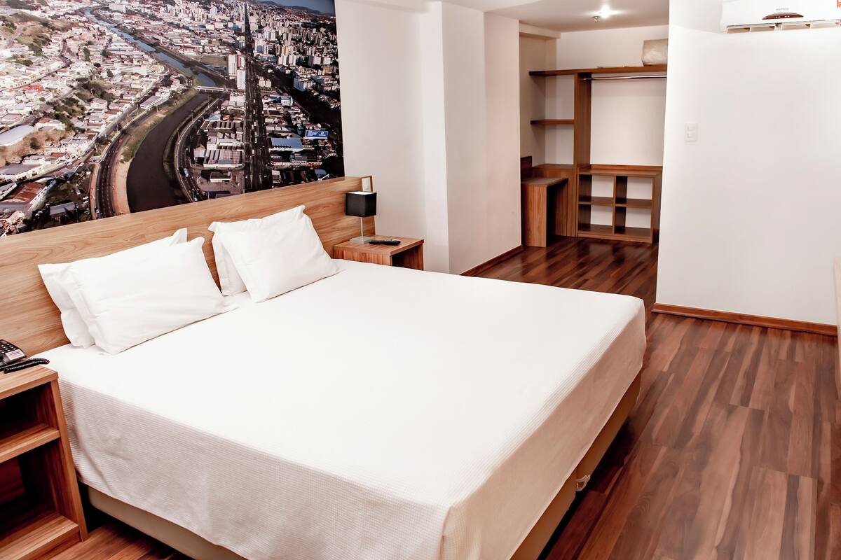 A hotel room with a large bed, wooden flooring, a nightstand with a phone, a lamp, and a view of a cityscape through the window.