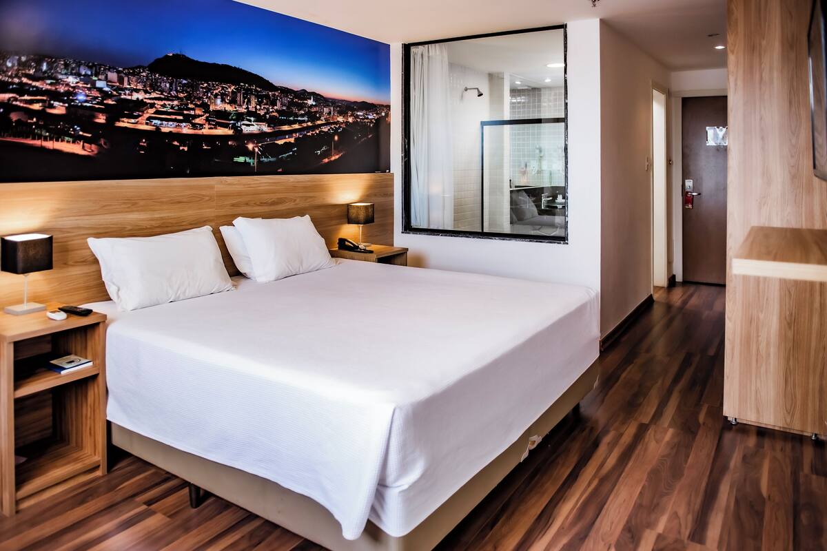 A hotel room with a large bed, wooden headboard, bedside tables with lamps, and a cityscape view on the wall.
