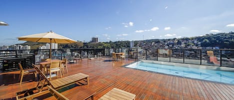 A rooftop pool area with wooden deck, lounge chairs, and a large umbrella.