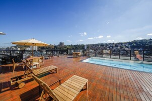 Área de piscina no terraço com deck de madeira, espreguiçadeiras e um grande guarda-sol.