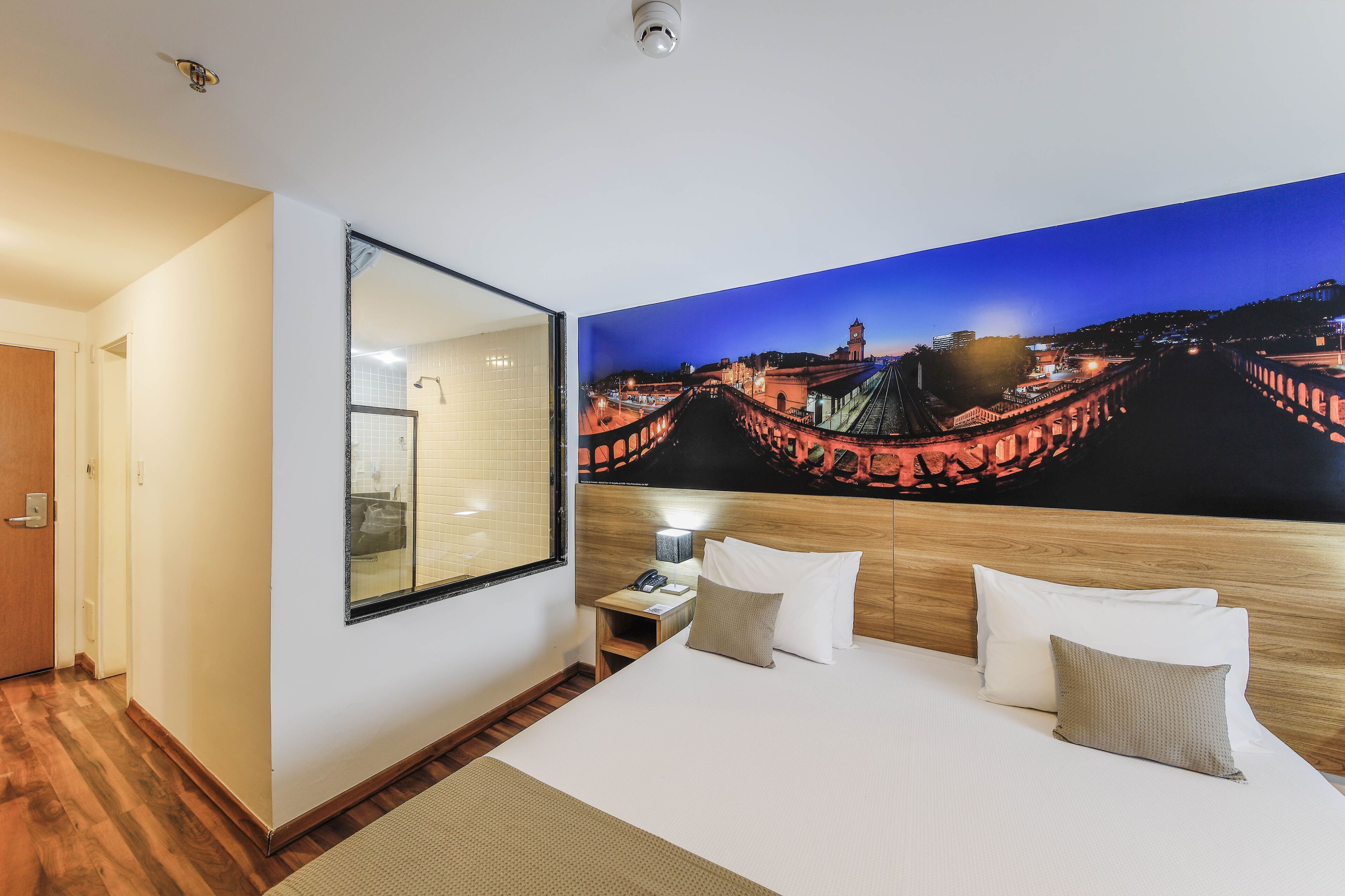A hotel room with a large bed, a nightstand, a mirror, and a wall-mounted TV displaying a cityscape.