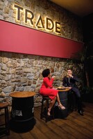 Two people seated at a table in a room with a stone wall and a 'TRADE' sign.
