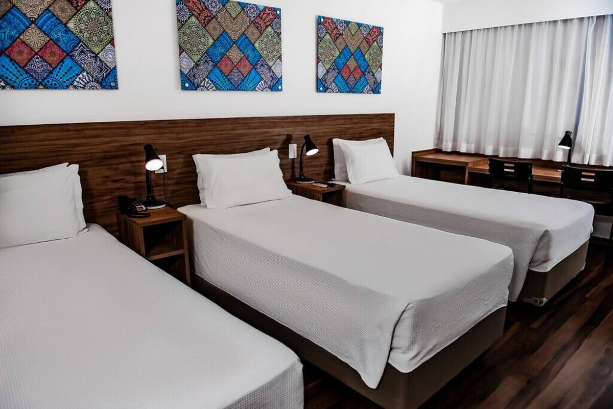 A hotel room with three single beds, a wooden headboard, bedside tables with lamps, and a desk with chairs.