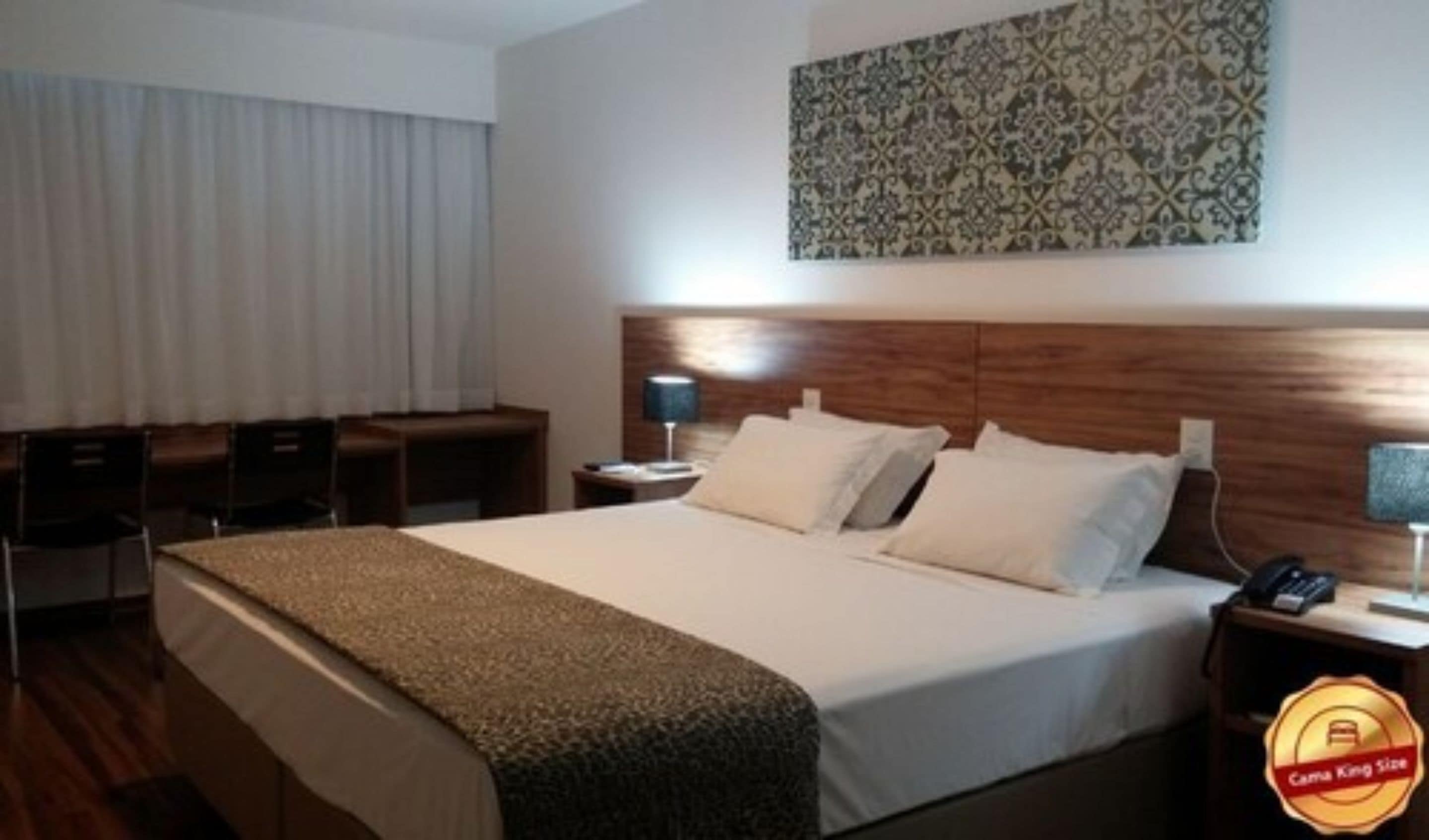 A hotel room with a large bed, a desk, a chair, and a nightstand with a lamp.