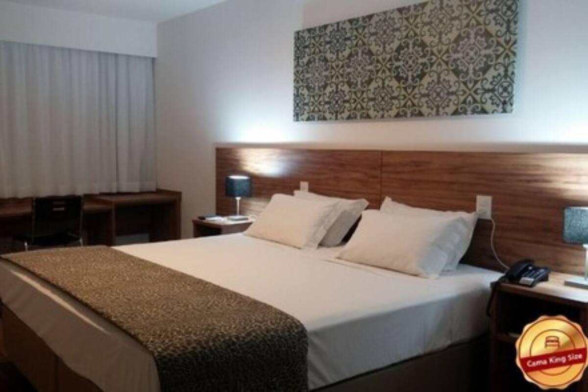 A hotel room with a large bed, a desk, a chair, and a nightstand with a lamp.