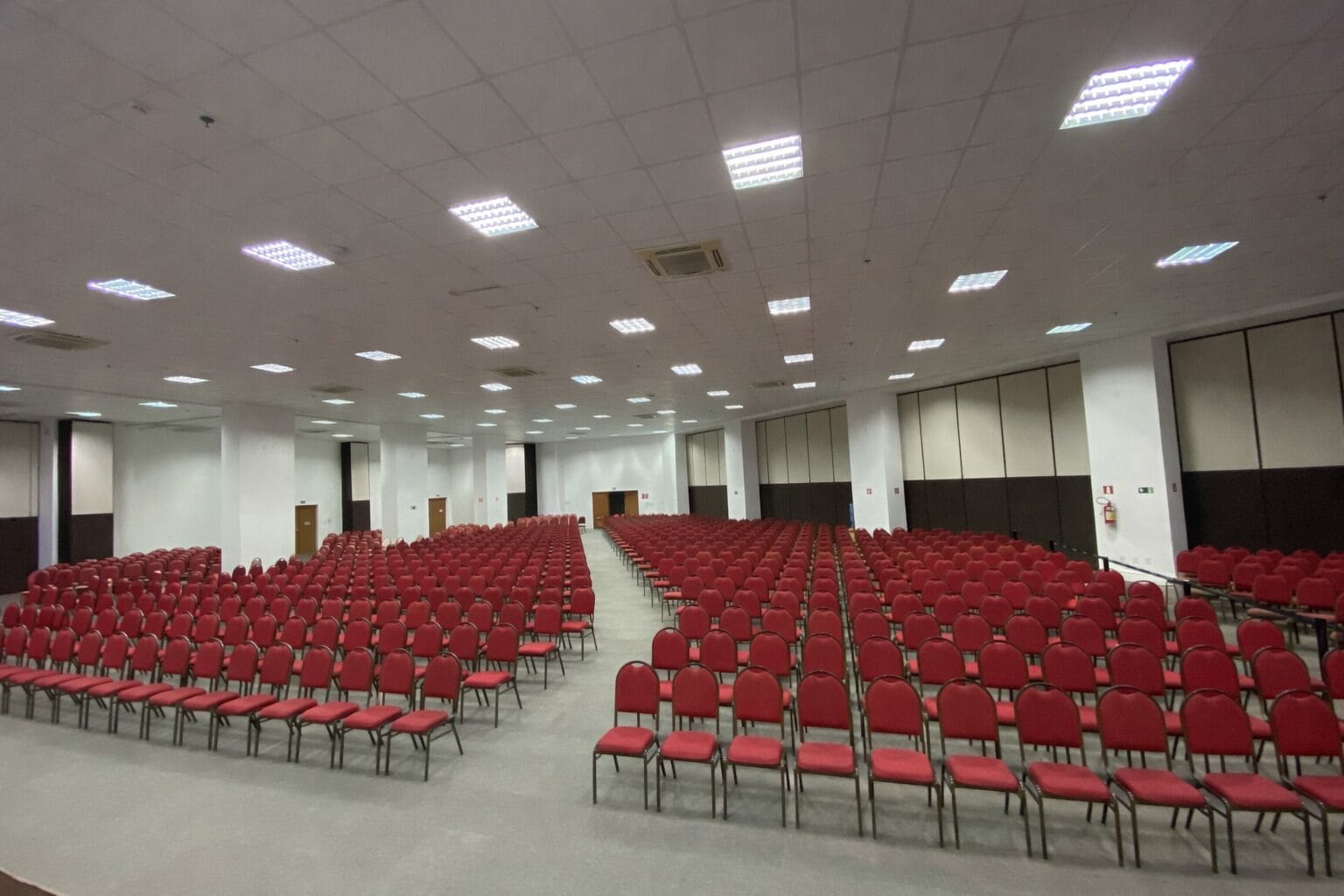 Un amplio salón de conferencias con filas de sillas rojas, un escenario frontal y un techo alto con iluminación fluorescente.