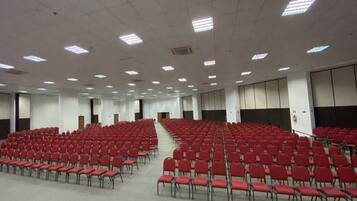 Un amplio salón de conferencias con filas de sillas rojas, un escenario frontal y un techo alto con iluminación fluorescente.