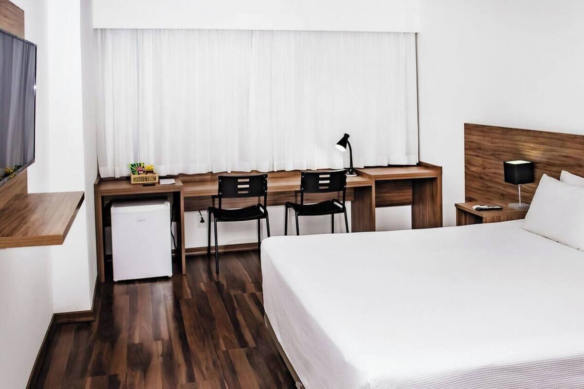 A hotel room with a bed, desk, chairs, a small refrigerator, and a TV.