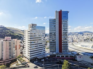 A hotel with a prominent red vertical stripe, surrounded by a cityscape with other buildings and a hilly background. - Trade Hotel (Juiz de Fora)