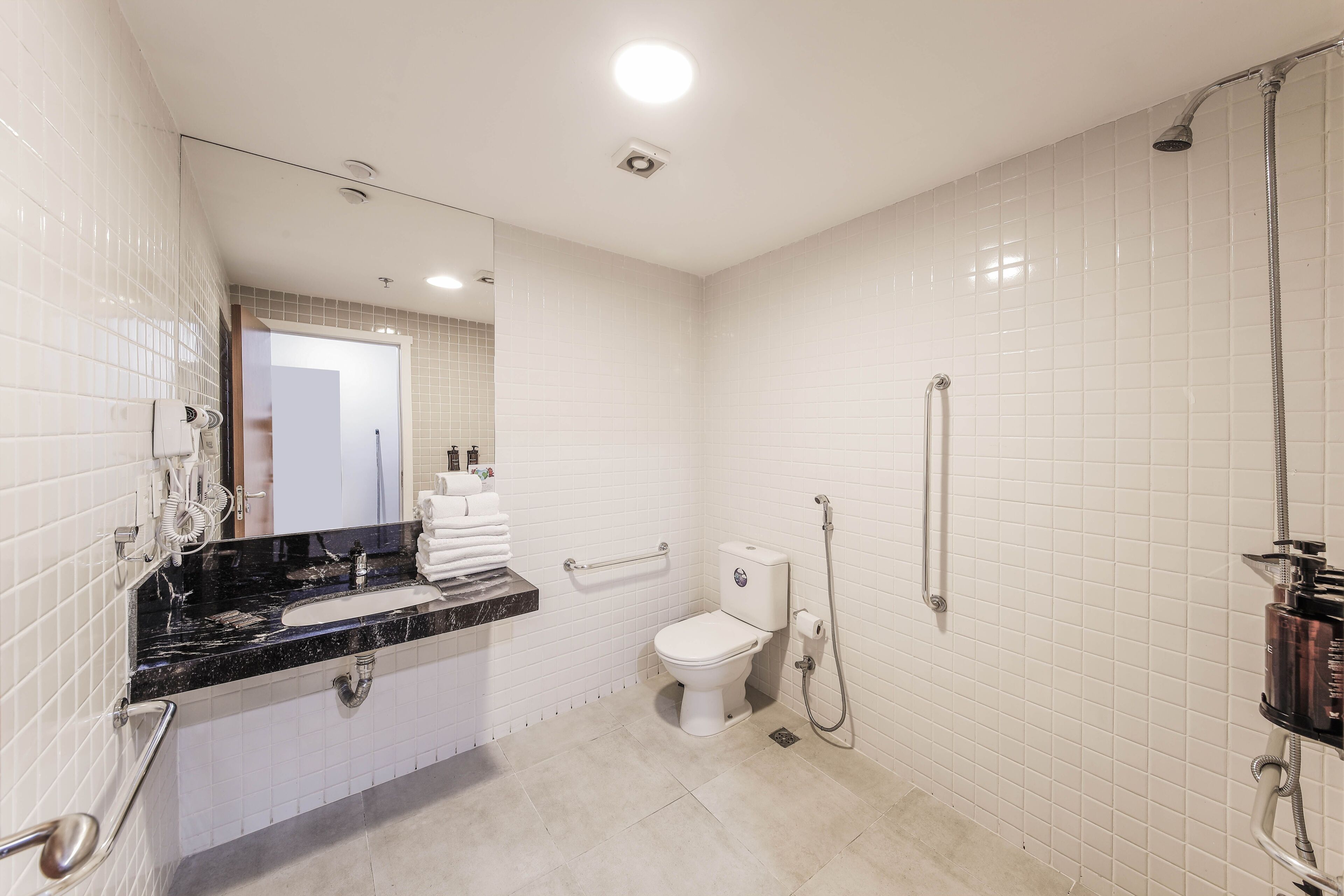 A bathroom with white tiled walls, a black marble sink, a toilet, a shower with a handheld hose, and a towel rack with folded towels.