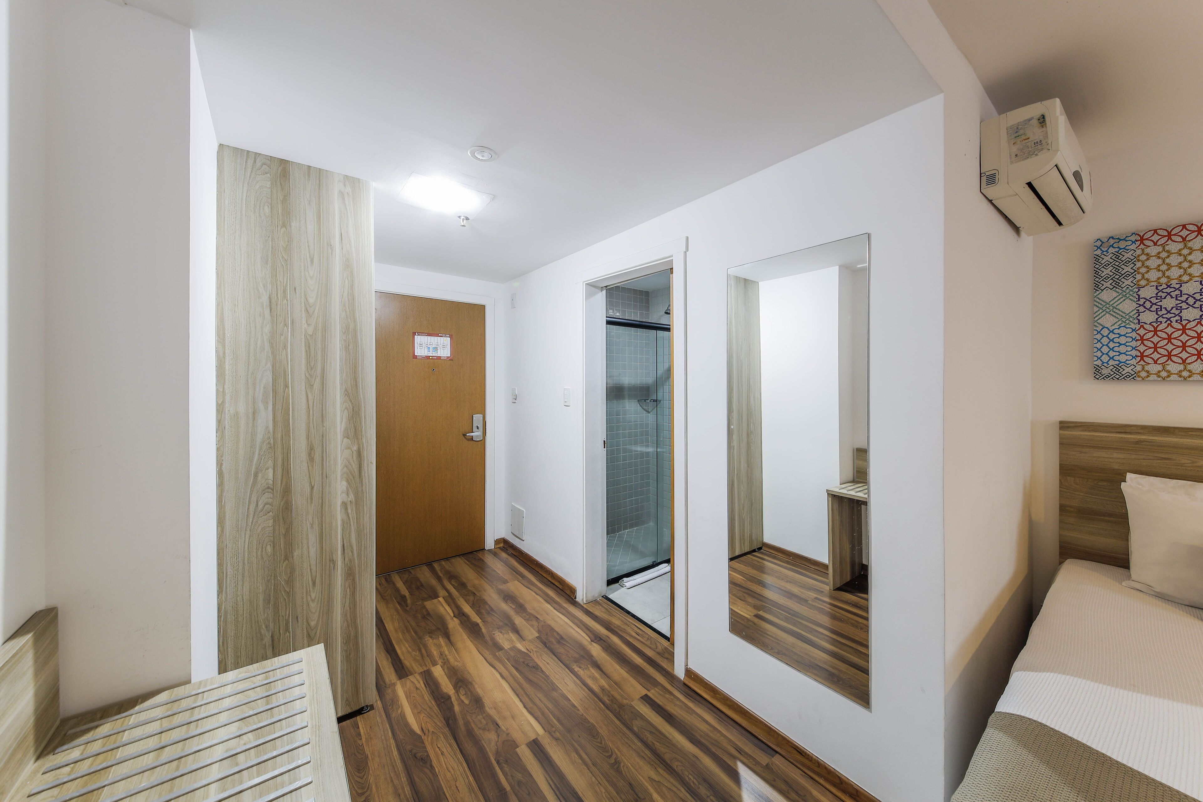 A hotel room with a wooden floor, a bed with a headboard, a large mirror, and a shower area.
