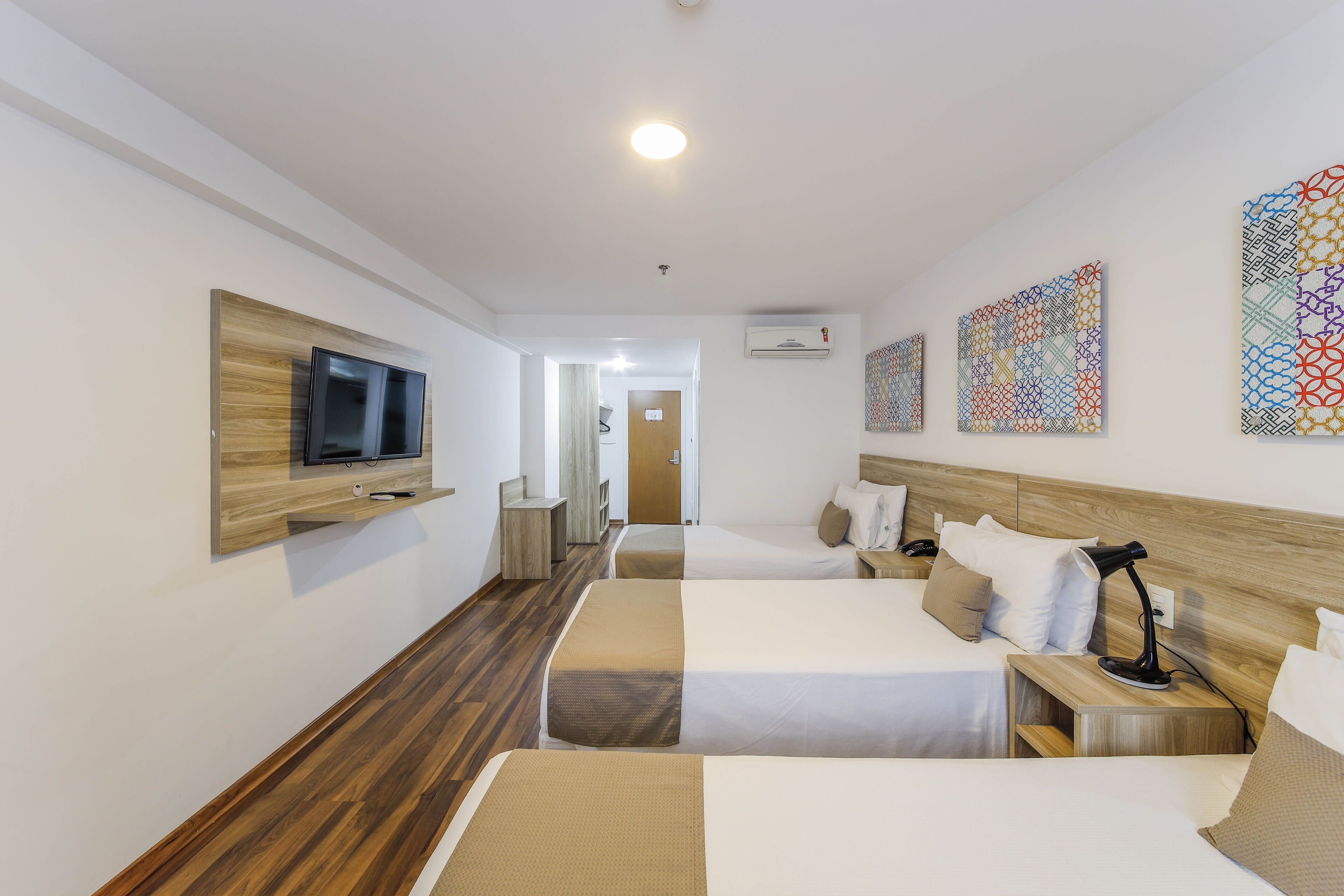 A hotel room with two beds, a TV mounted on a wooden panel, an air conditioning unit, and decorative wall art.