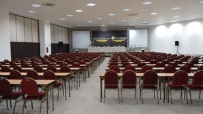 A conference room with rows of desks and chairs, a stage with a presentation screen, and a banner indicating an event. - Trade Hotel (Juiz de Fora)