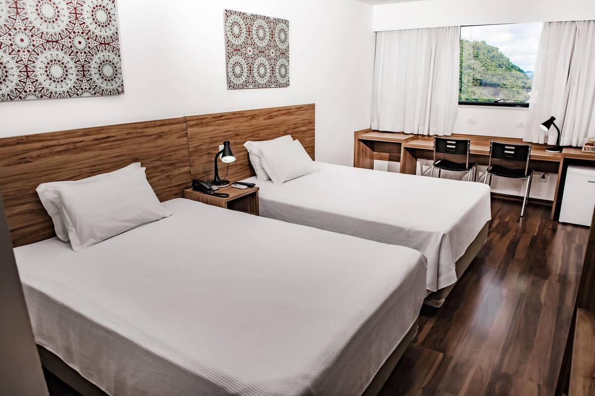 A hotel room with two beds, a desk with chairs, a small kitchenette, and a window with a view of greenery.