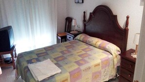 Een slaapkamer met een houten bed, een nachtkastje, een stoel, een televisie en een raam met gordijnen.