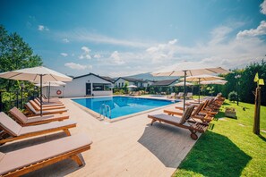 Ein Schwimmbecken mit Liegestühlen und Sonnenschirmen, umgeben von Grünflächen und einem klaren Himmel.