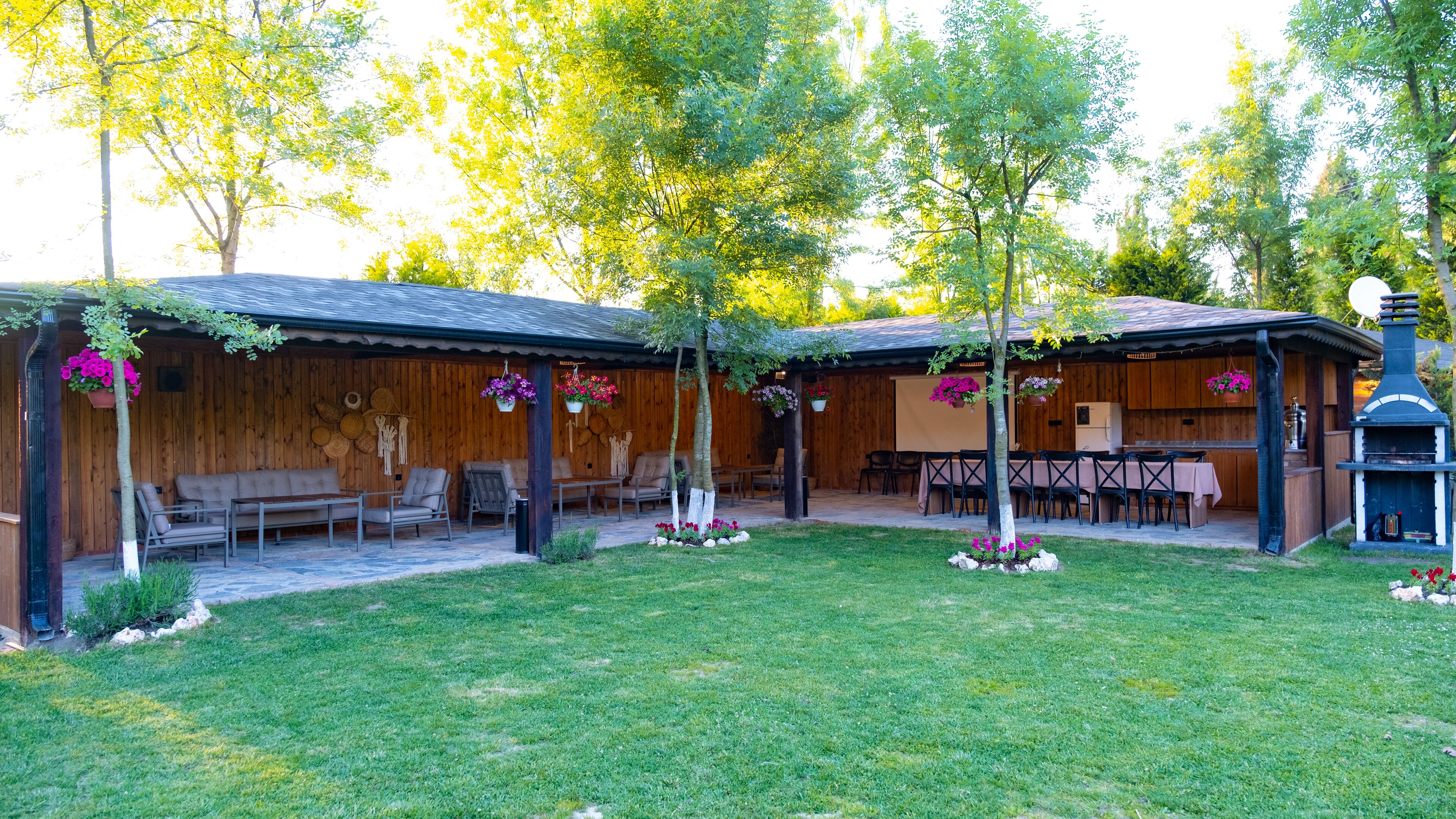 Une terrasse extérieure rustique, avec des bancs et des tables en bois, entourée d’une pelouse bien entretenue et d’arbres.