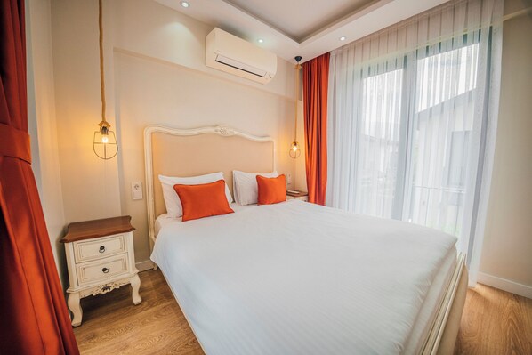 A neatly made bed with white and orange bedding, a wooden nightstand with drawers, and a window with sheer curtains.