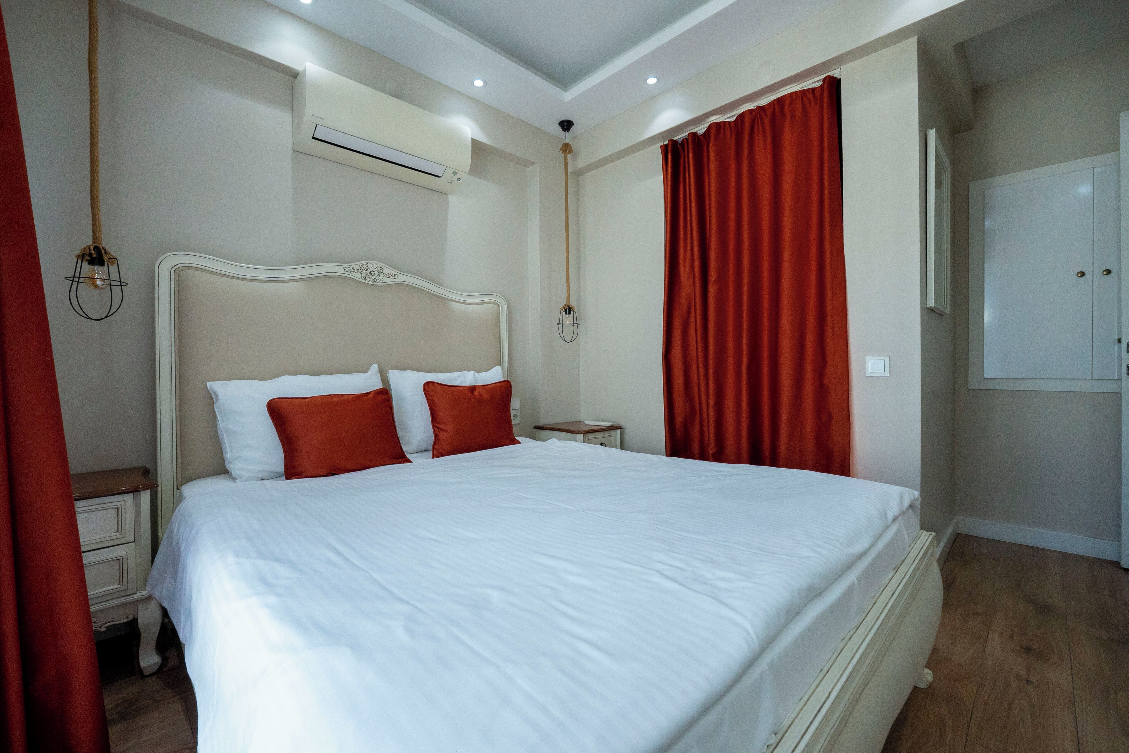 A neatly made bed with white and red bedding, a bedside table, an air conditioning unit, and a wardrobe in a hotel room.