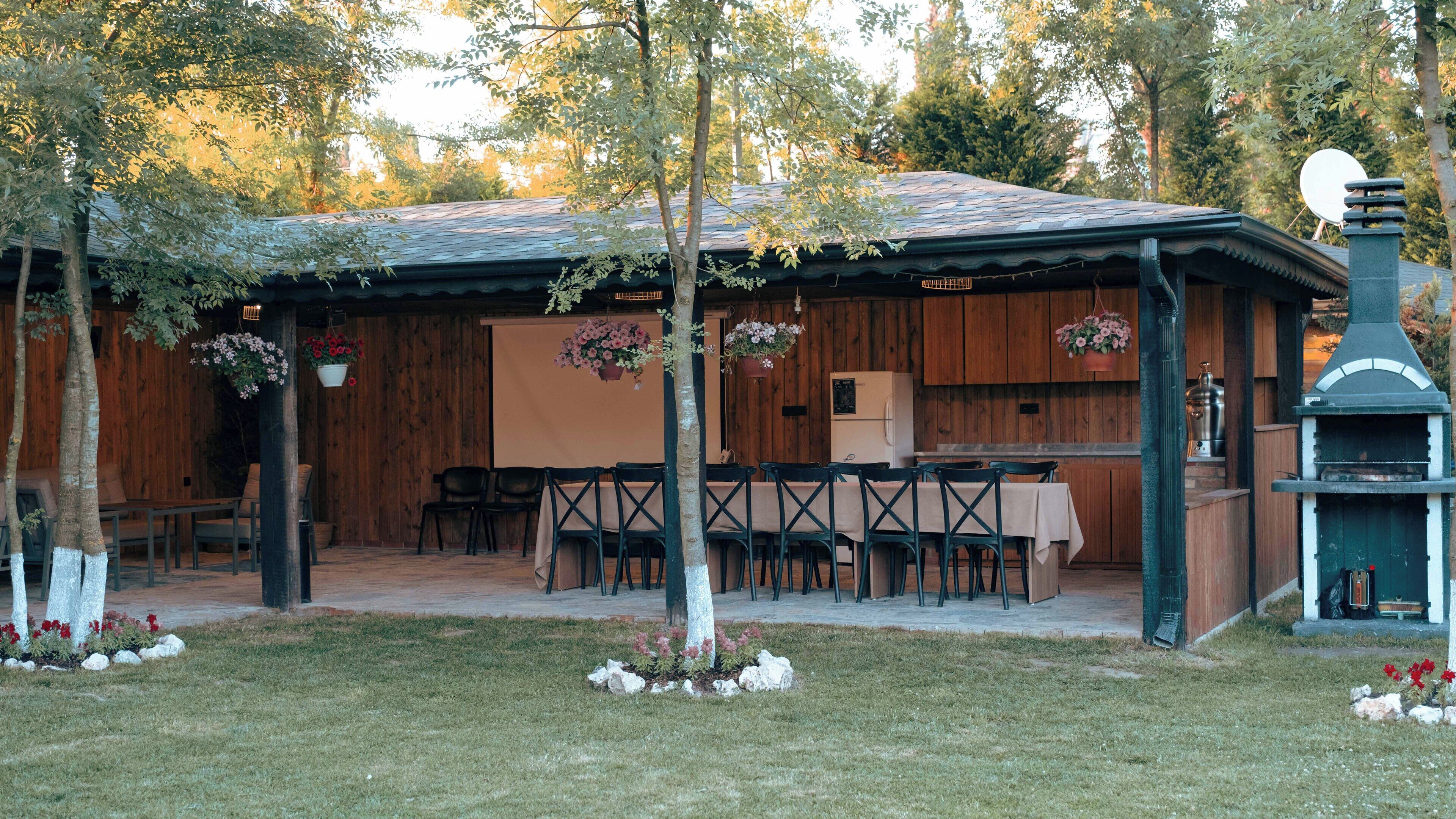 A rustic outdoor dining area with wooden tables and chairs, a kitchen area with wooden cabinets, and a chimney.