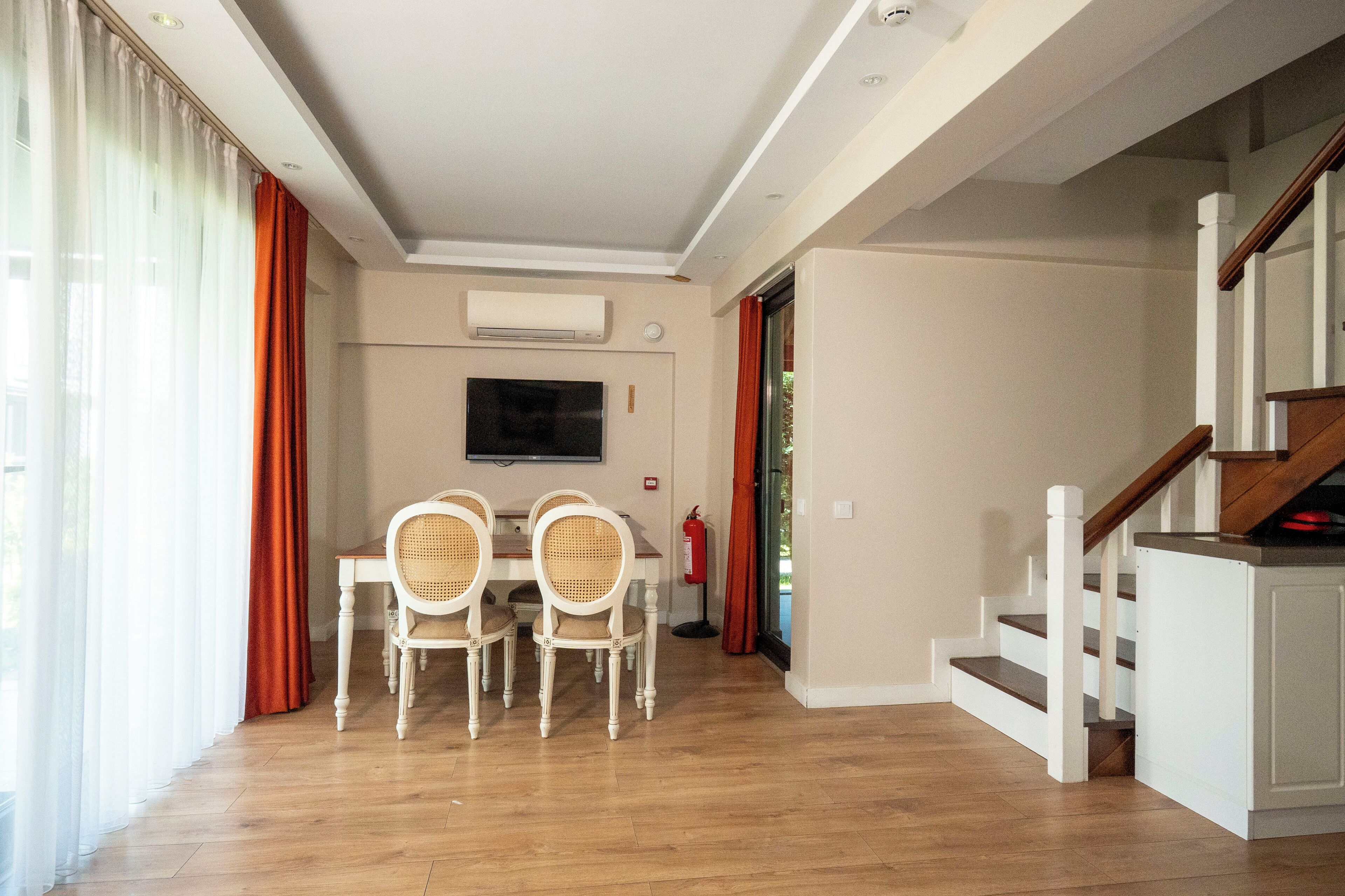 A spacious living area with a dining table and chairs, a flat-screen TV mounted on the wall, and a staircase leading to an upper level.