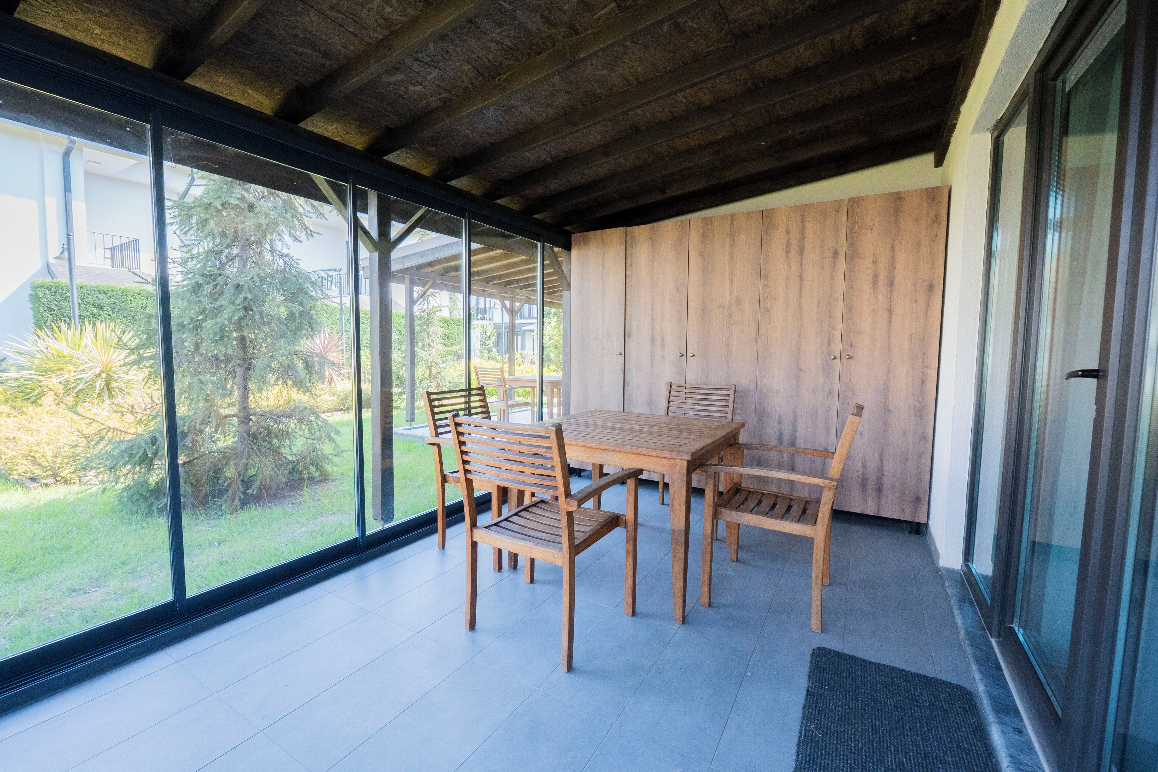 A modern patio with a wooden table and chairs, large windows, and a view of greenery.