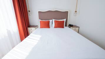 Un letto rifatto con cura, lenzuola bianche e due cuscini arancioni, una testiera in legno e due comodini con lampade.