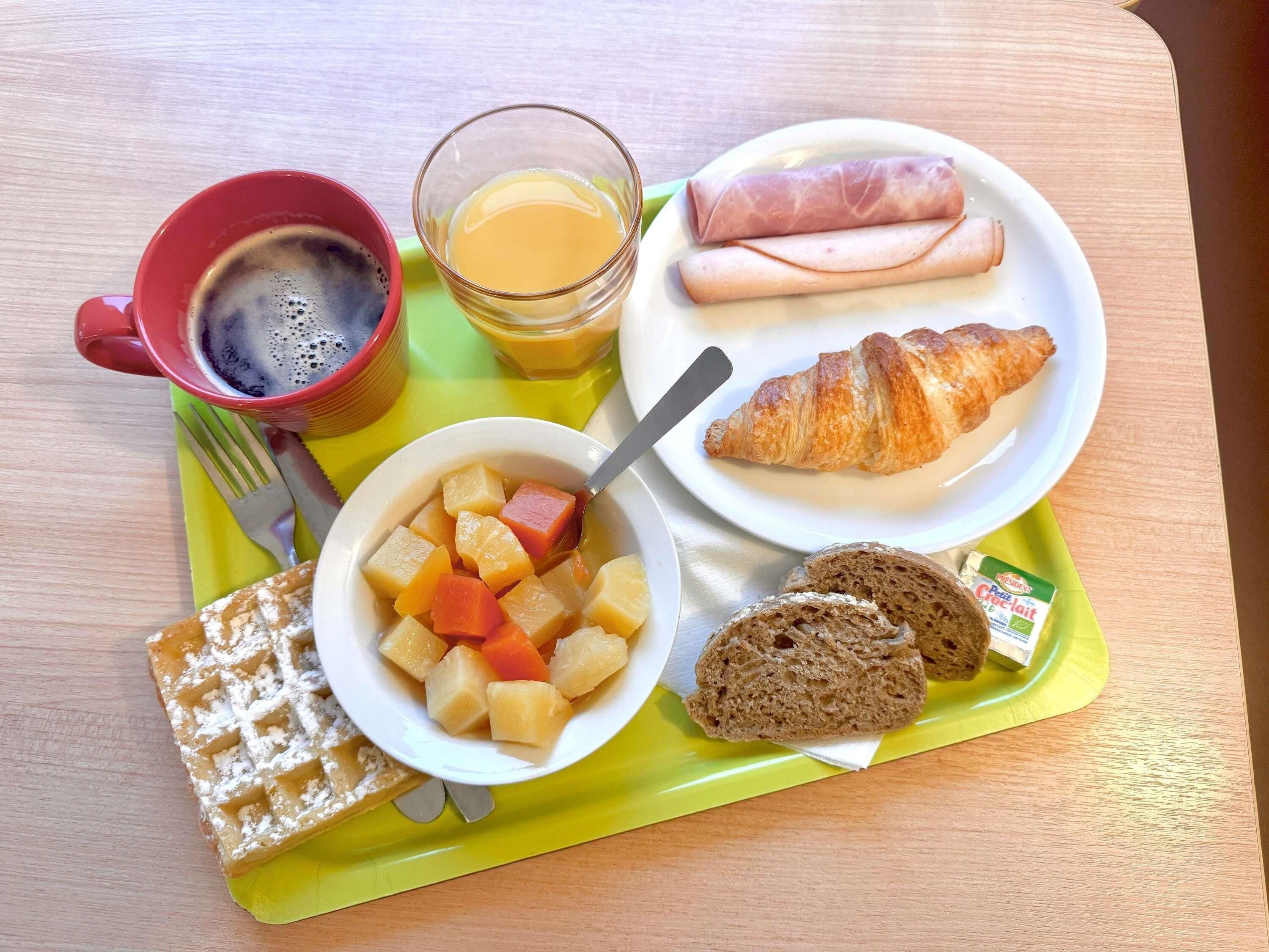 Een dienblad met ontbijtartikelen, waaronder wafels, fruit, croissants en een warm drankje.