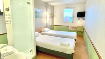 Una habitación de hotel pequeña con dos camas individuales, un baño con lavamanos e inodoro, un televisor montado en la pared y una ventana con vista al cielo.
