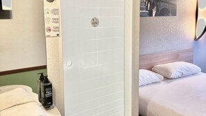 A hotel room with a glass shower door, a sink with a towel, a bottle of shampoo, and a bed with pillows. - ibis budget Cosne-sur-Loire (Cosne-Cours-sur-Loire)