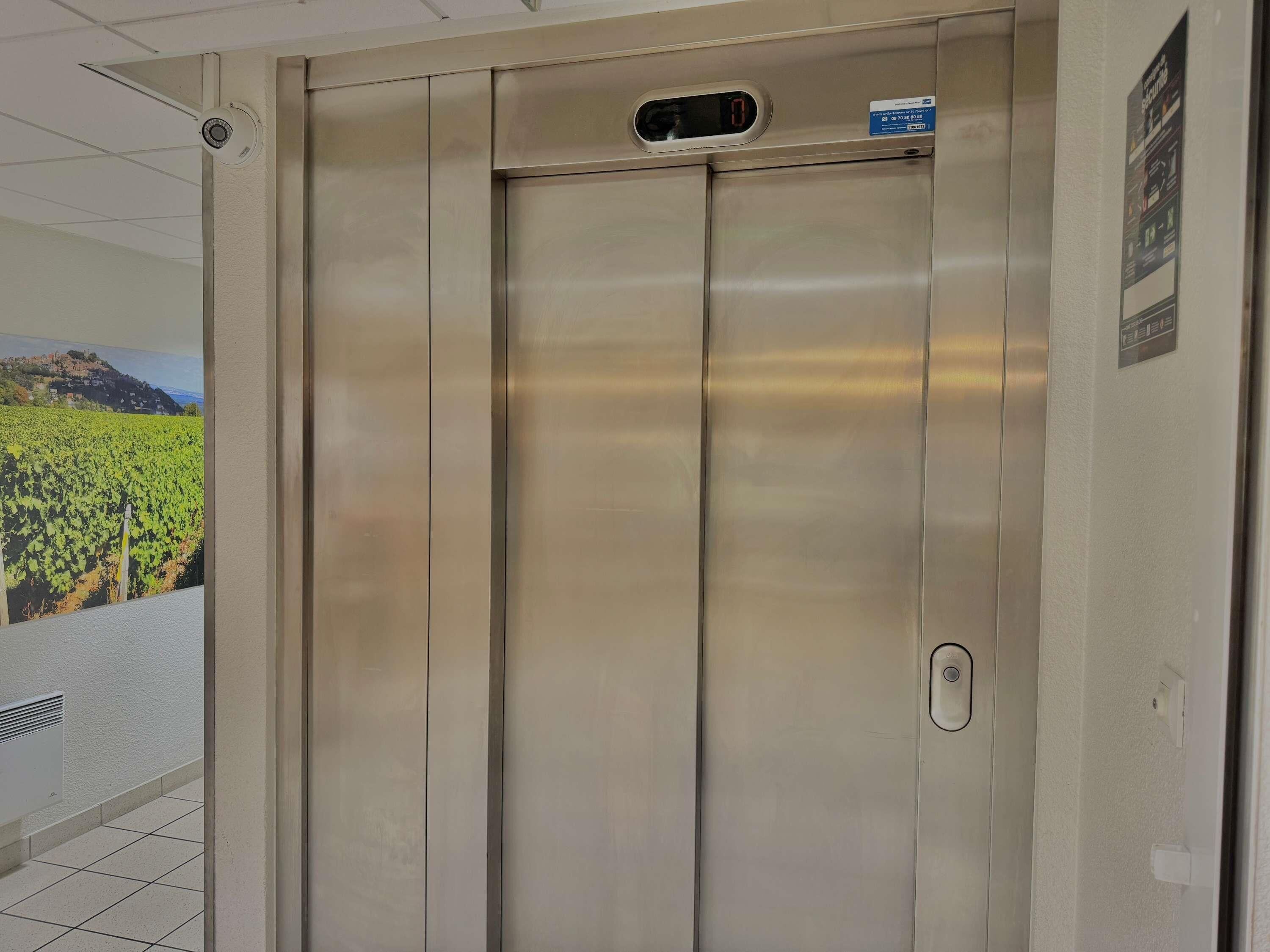 A metallic elevator with a digital display, a vent, and a poster on the wall.