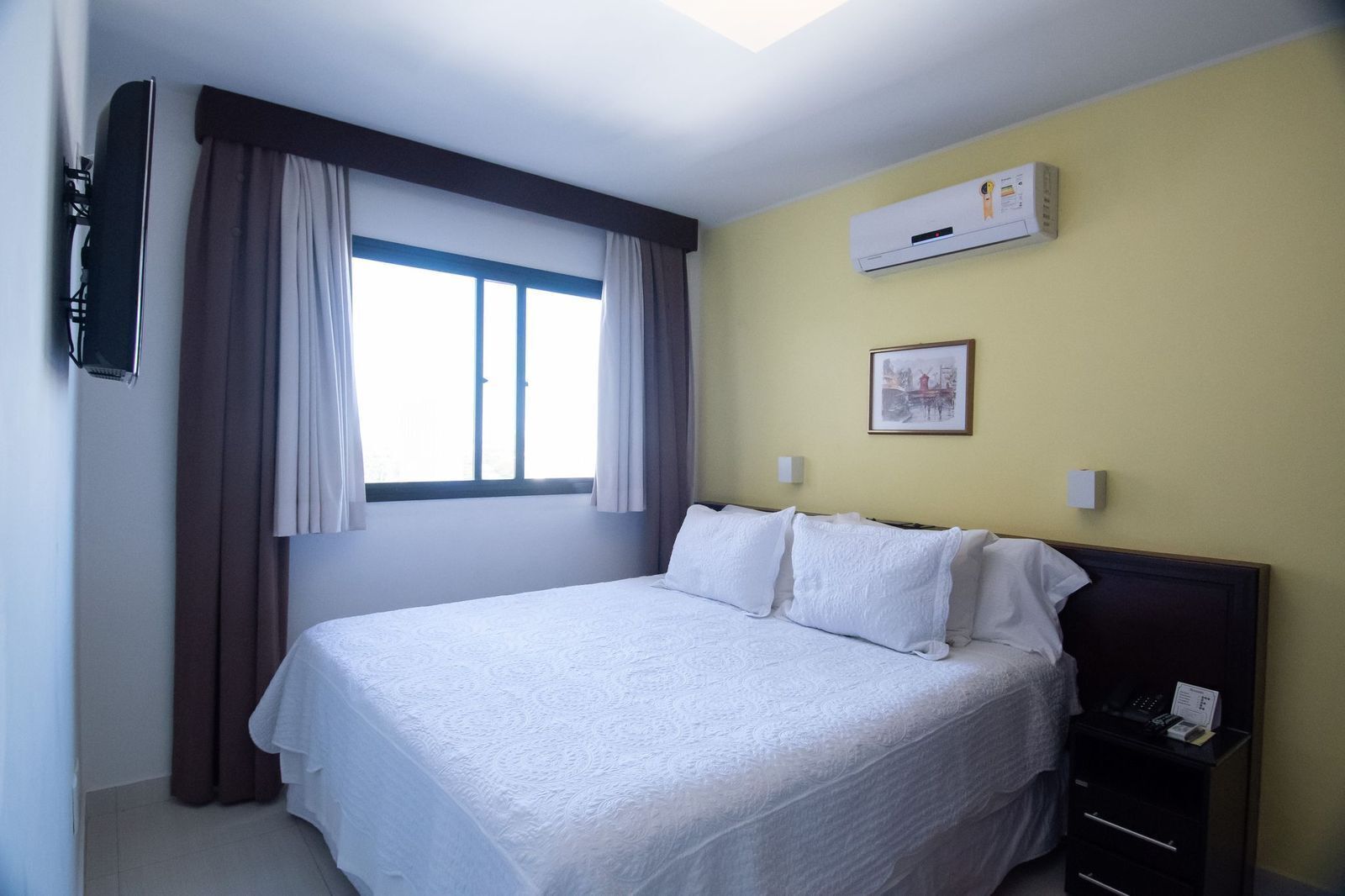 A hotel room with a bed, a bedside table, an air conditioning unit, a window with curtains, and a wall-mounted picture.
