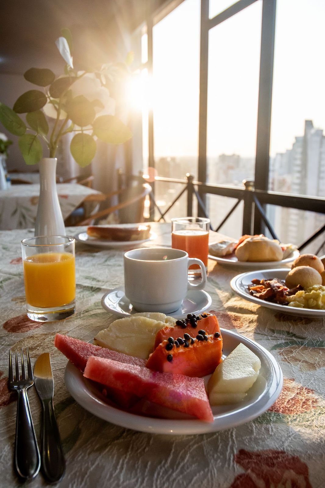 Een ontbijttafel met fruit, toast en eieren, een glas sinaasappelsap en een kop koffie, met uitzicht op een stadsgezicht.